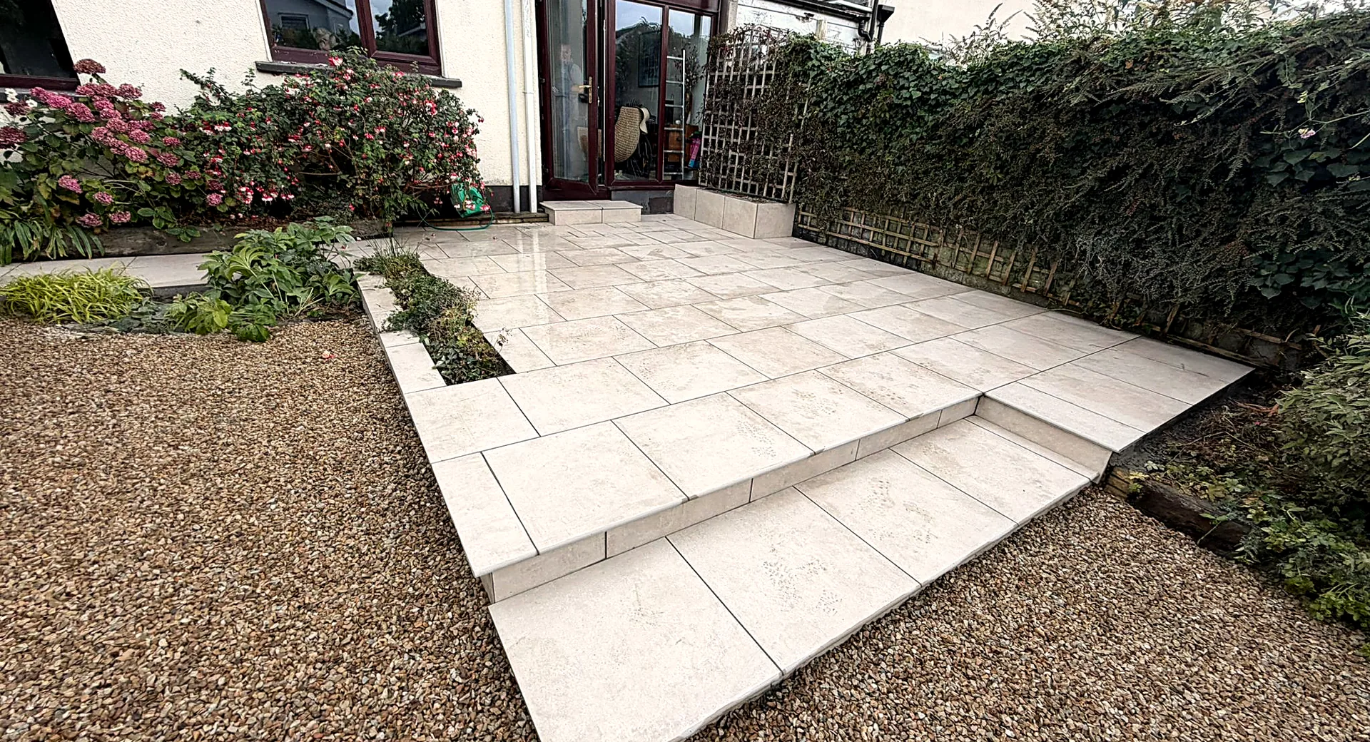Finished large-format porcelain patio at the glazed back door of the Stillorgan house, with a step down to the lower level, a planted bed beside the wall and gravel borders