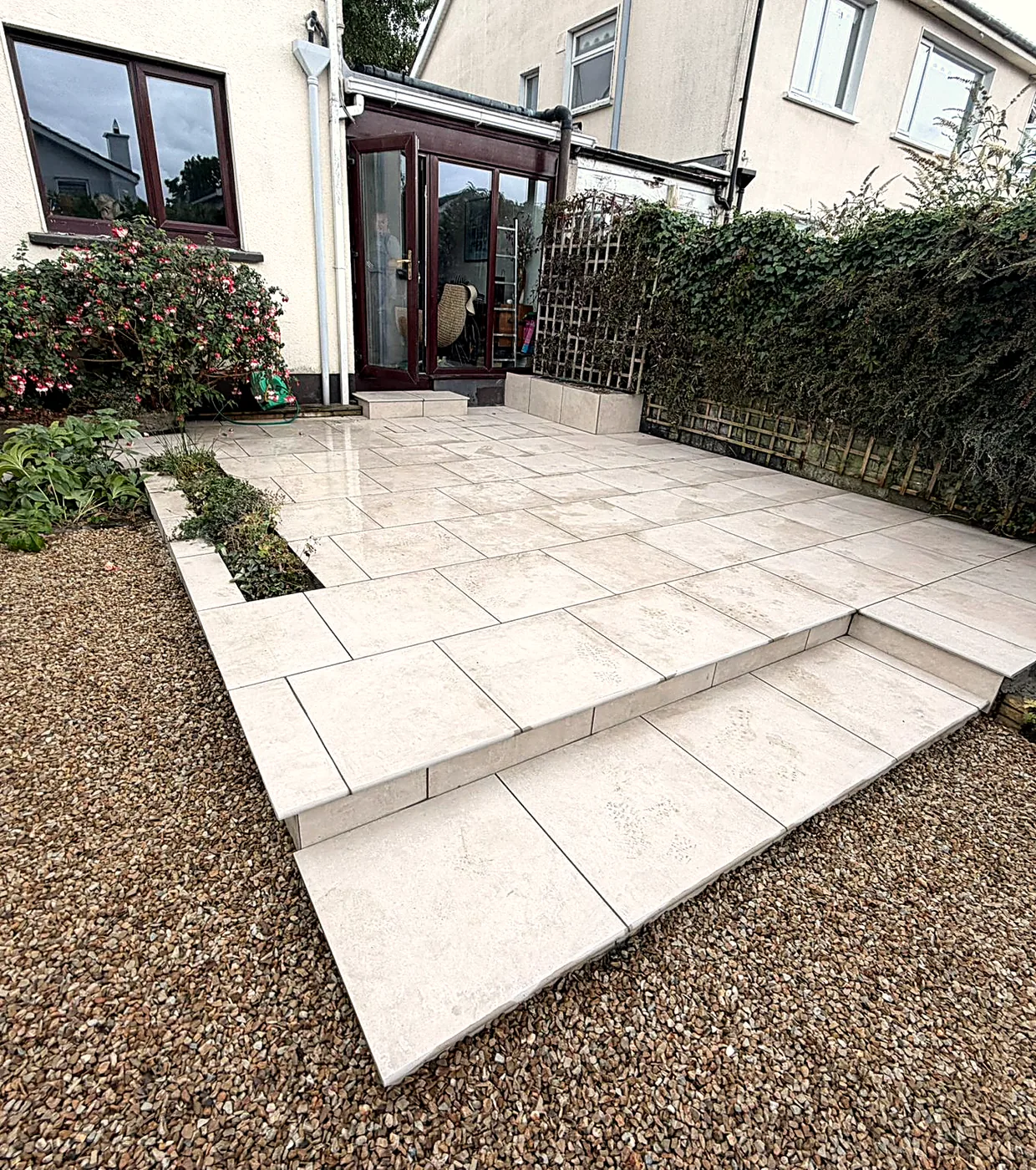 Finished large-format porcelain patio at the glazed back door of the Stillorgan house, with a step down to the lower level, a planted bed beside the wall and gravel borders