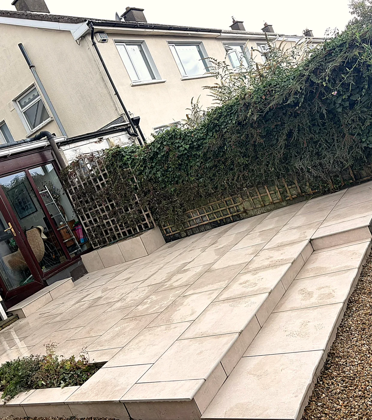 The Stillorgan patio from the garden: two levels of cream porcelain with a long step between them, the house behind and a tall hedge along the boundary
